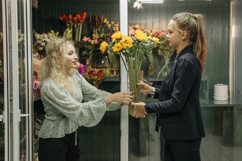 A florist with curly blonde hair, wearing a light green floral blouse, is handing a bouquet of yellow sunflowers to a customer with long brown hair tied back, dressed in a dark blazer, inside a floral