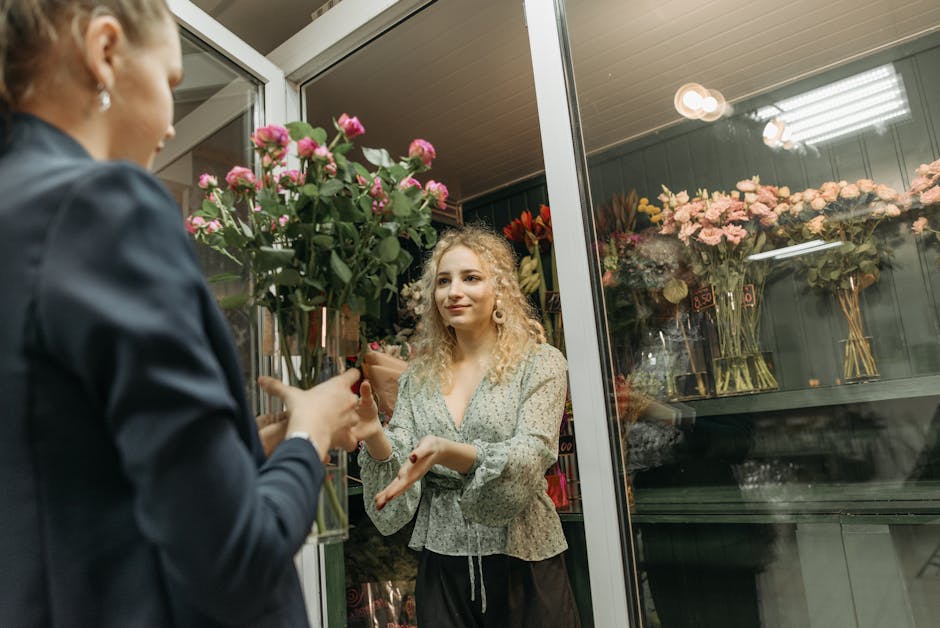 A woman with curly blonde hair and wearing a light patterned blouse is inside a flower shop, receiving a bouquet of pink roses from a florist whose back is partially visible. The bouquet is arranged w