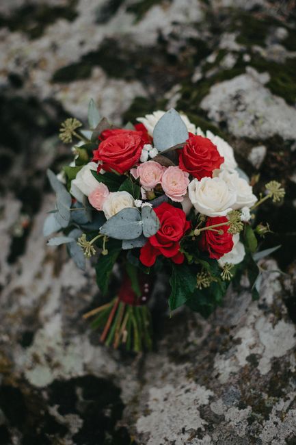 A bouquet of fresh flowers resting on a textured stone surface, featuring red roses, white roses, and pale pink ranunculus blossoms. The arrangement includes dusty miller leaves with soft, silvery-gre